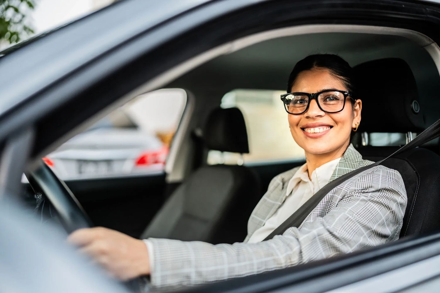 Smiling woman wearing glasses driving a car with a seatbelt on.