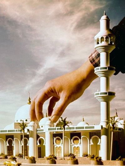 A hand adjusting a detailed miniature mosque model under a cloudy sky.