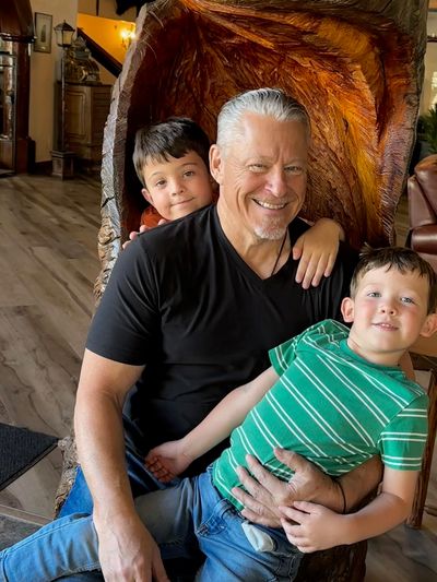 Man with two young boys sitting on his lap, smiling.