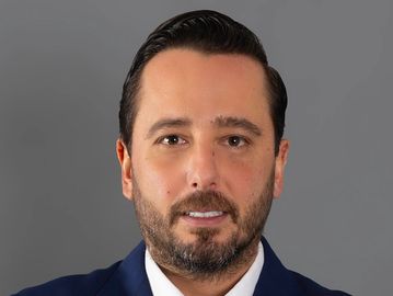 Professional headshot of a man with dark hair and beard in a suit.