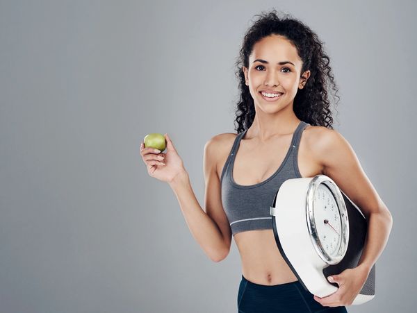 A fit female person holding an apple and a weighing machine in her hand