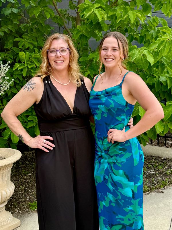 Two women smiling outdoors, dressed in elegant dresses, standing in front of green foliage.