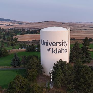 Large water tower with "University of Idaho" text surrounded by trees and hills.