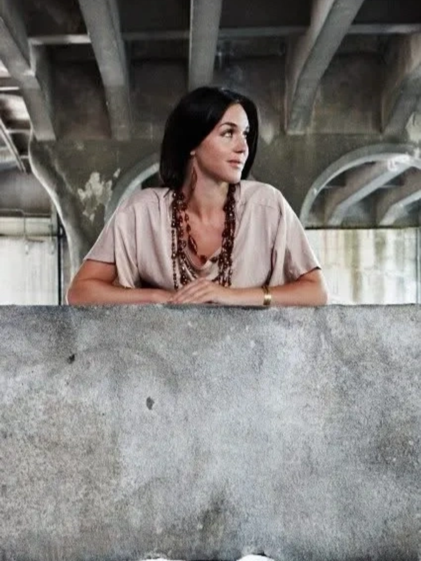 Woman leaning on concrete wall under a bridge, looking sideways.