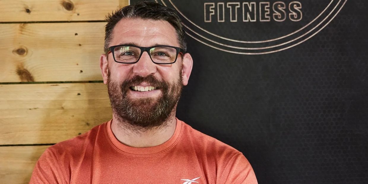 Smiling man in glasses and orange shirt standing in front of a fitness sign.