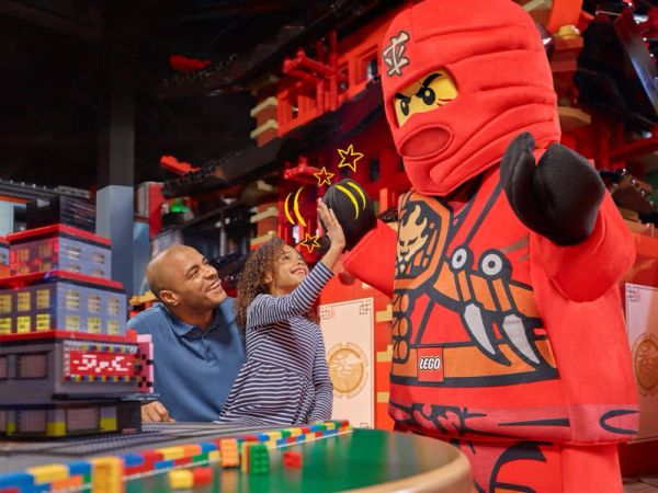 Child high-fives LEGO ninja character with dad watching.