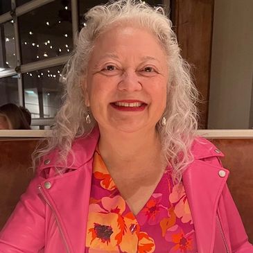 Photo of smiling, brown-eyed woman with curly, grey hair wearing a bright pink jacket and blouse. 