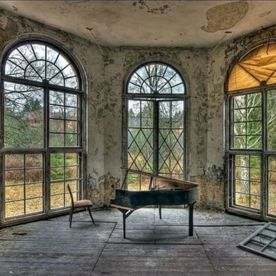 An old piano and chair in a dilapidated room with large arched windows.