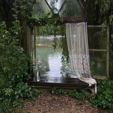 Rustic window with lace curtain overlooking a tranquil lake surrounded by greenery.