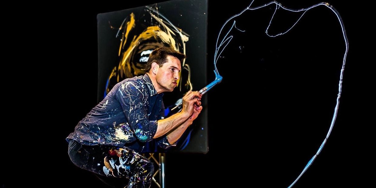 man crouching with a stand of paint coming from paintbrushes in his hands