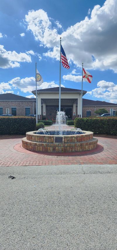 Fountain with three flags in front of a brick building under a blue sky with clouds.