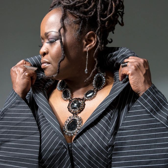 Woman with pin-stripe top and giant bejeweled necklace on her bared neck. She's grasping her lapels.