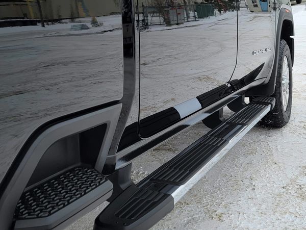 Close-up of a black vehicle's side step and tire on snowy ground.