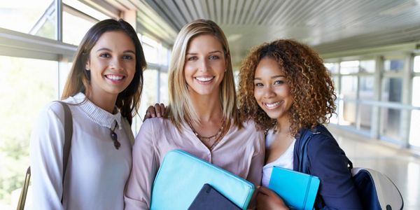 Three smiling female students holding folders and backpacks in a school hallway.
