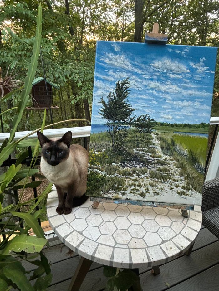 A Siamese cat next to a landscape painting on a tiled table outdoors.
