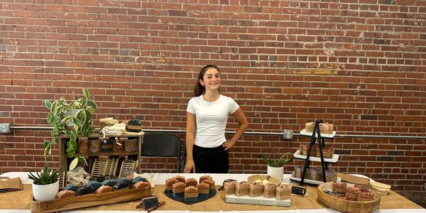 Young woman selling assorted baked goods at a market stall with brick wall backdrop.