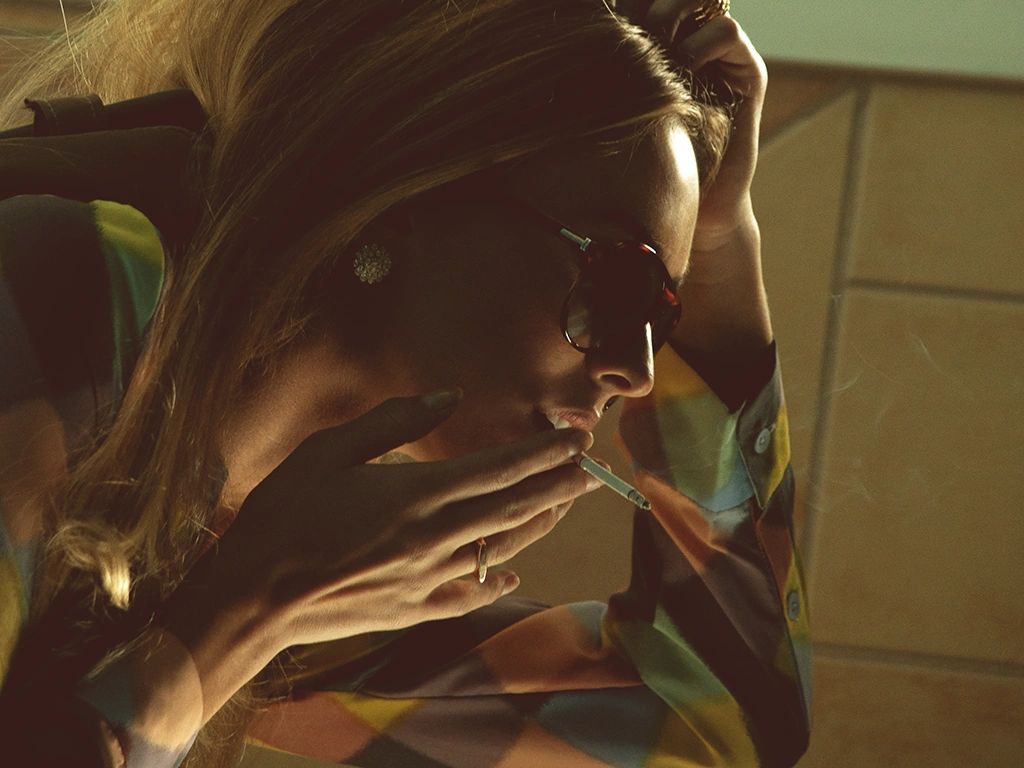 Woman wearing sunglasses smoking a cigarette indoors.