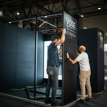 Two men assembling a black exhibition booth with the sign 'VOSE'.