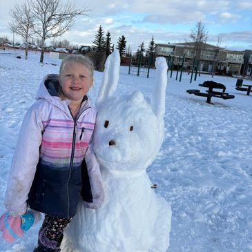 A girl smiles beside a bunny-shaped snow sculpture in a snowy park.