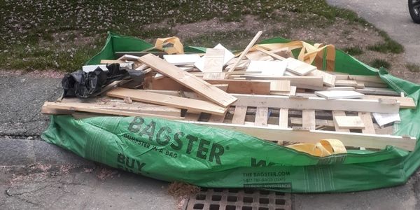 A green Bagster dumpster bag filled with wooden debris on pavement.
