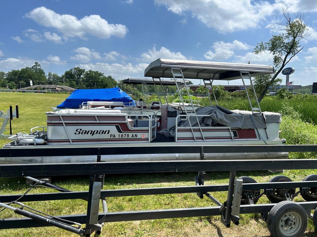 Pontoon boat parked on trailer near grassy field under blue sky.