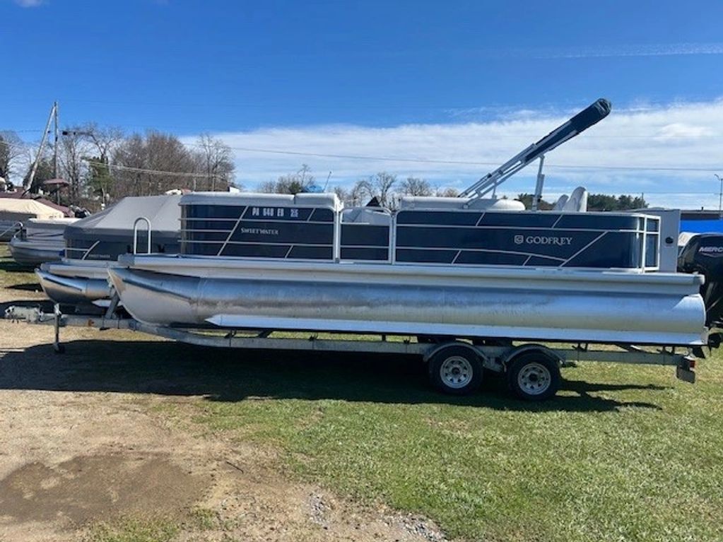 A Godfrey pontoon boat on a trailer parked on grass under a clear sky.
