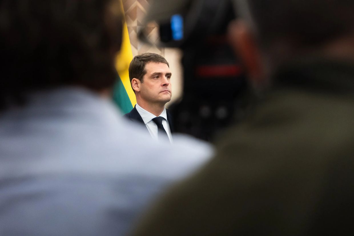 A serious man in a suit is seen through a crowd, with a flag in the background.