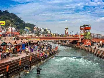 Crowded riverbank with people bathing and a bridge under a partly cloudy sky.