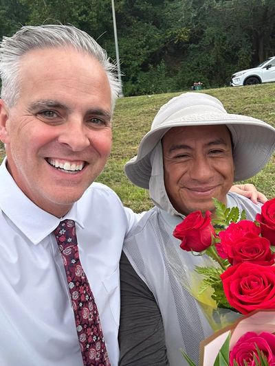 Two men smiling, one holding a bouquet of red roses outdoors.