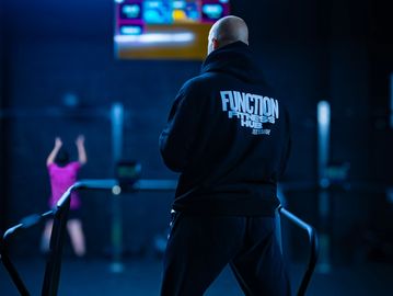 Man in a black hoodie at a gym with a woman exercising in the background.