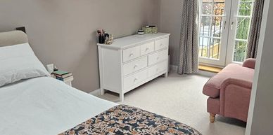 Cozy bedroom with floral bedspread, white dresser, and pink armchair near window.