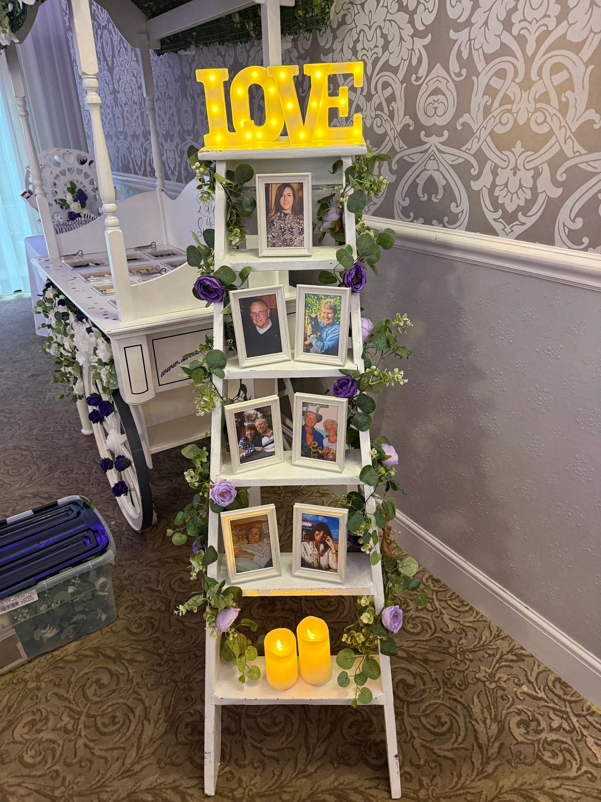 A white ladder shelf with framed photos, purple flowers, candles, and a lit LOVE sign.
