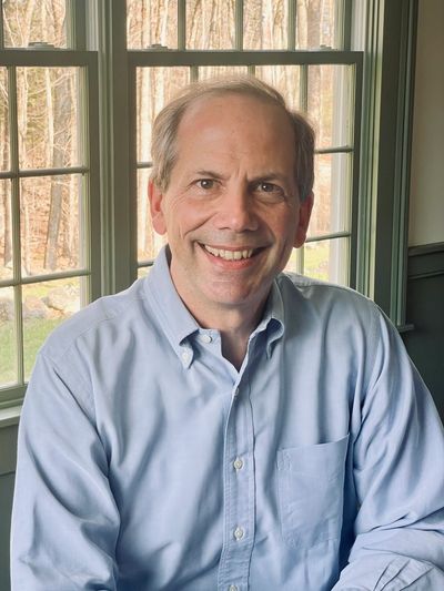 Smiling middle-aged man in a light blue shirt sitting by a window.