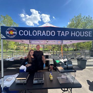 Two people stand under a Colorado Tap House tent with cooking equipment on a sunny day.