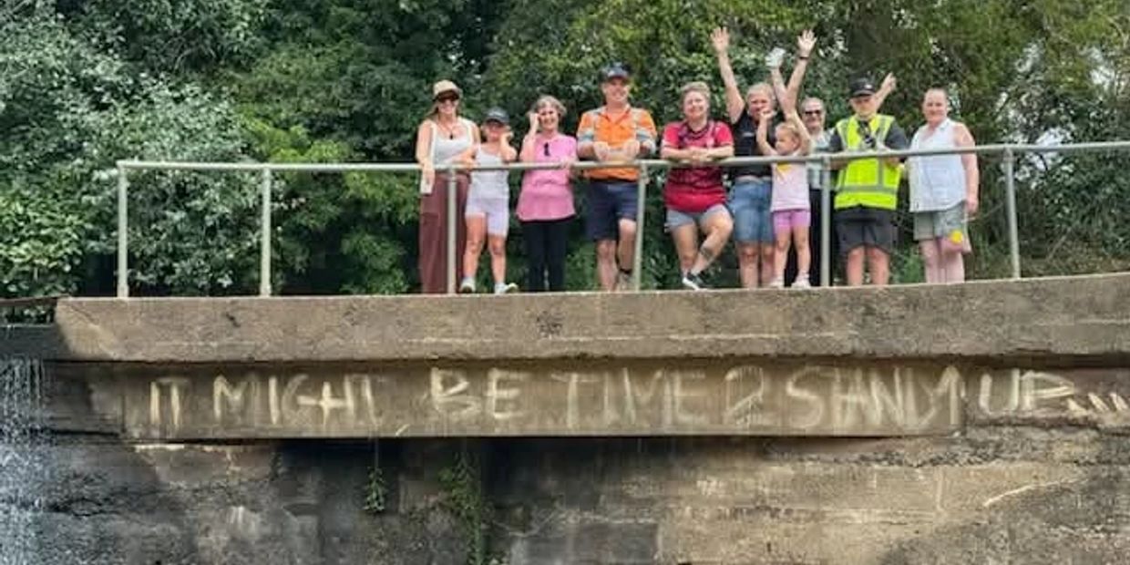 A group of people standing on a bridge with graffiti that reads 'It might be time 2 sand up'.