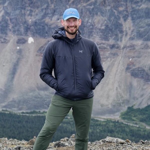 Smiling man in outdoor jacket and cap standing on rocky terrain with mountains behind.