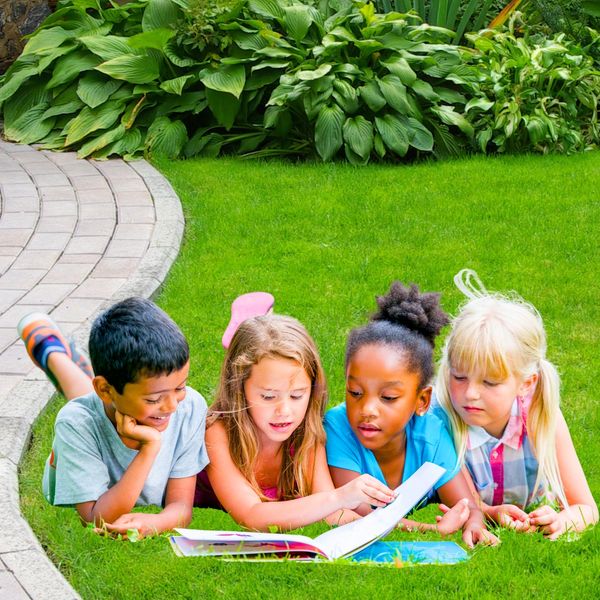 Four diverse children lying on grass reading a book together outdoors.