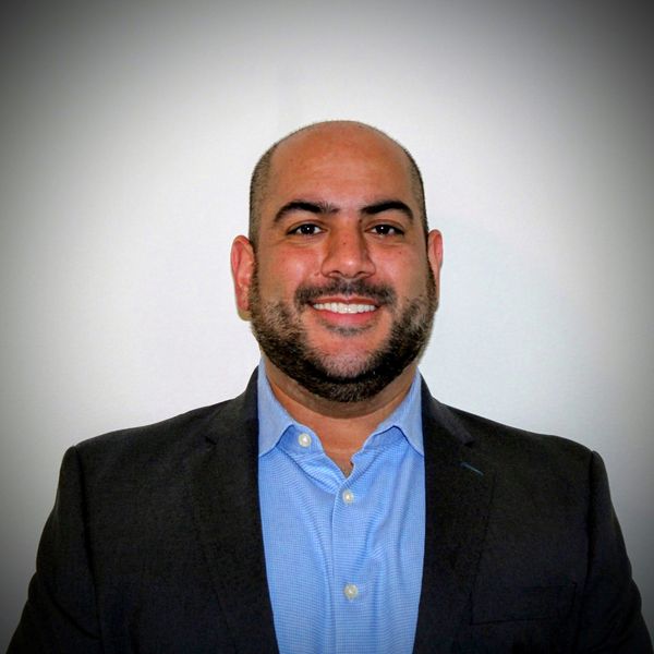 Smiling man in a blue shirt and dark blazer against a plain background.