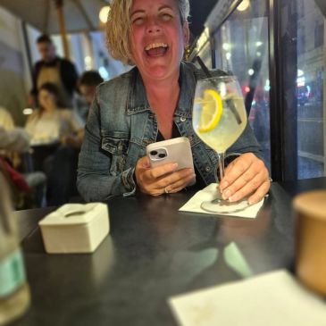 Woman laughing with a cocktail and phone at a table.