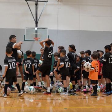Children in soccer uniforms gather around a coach in an indoor gym.