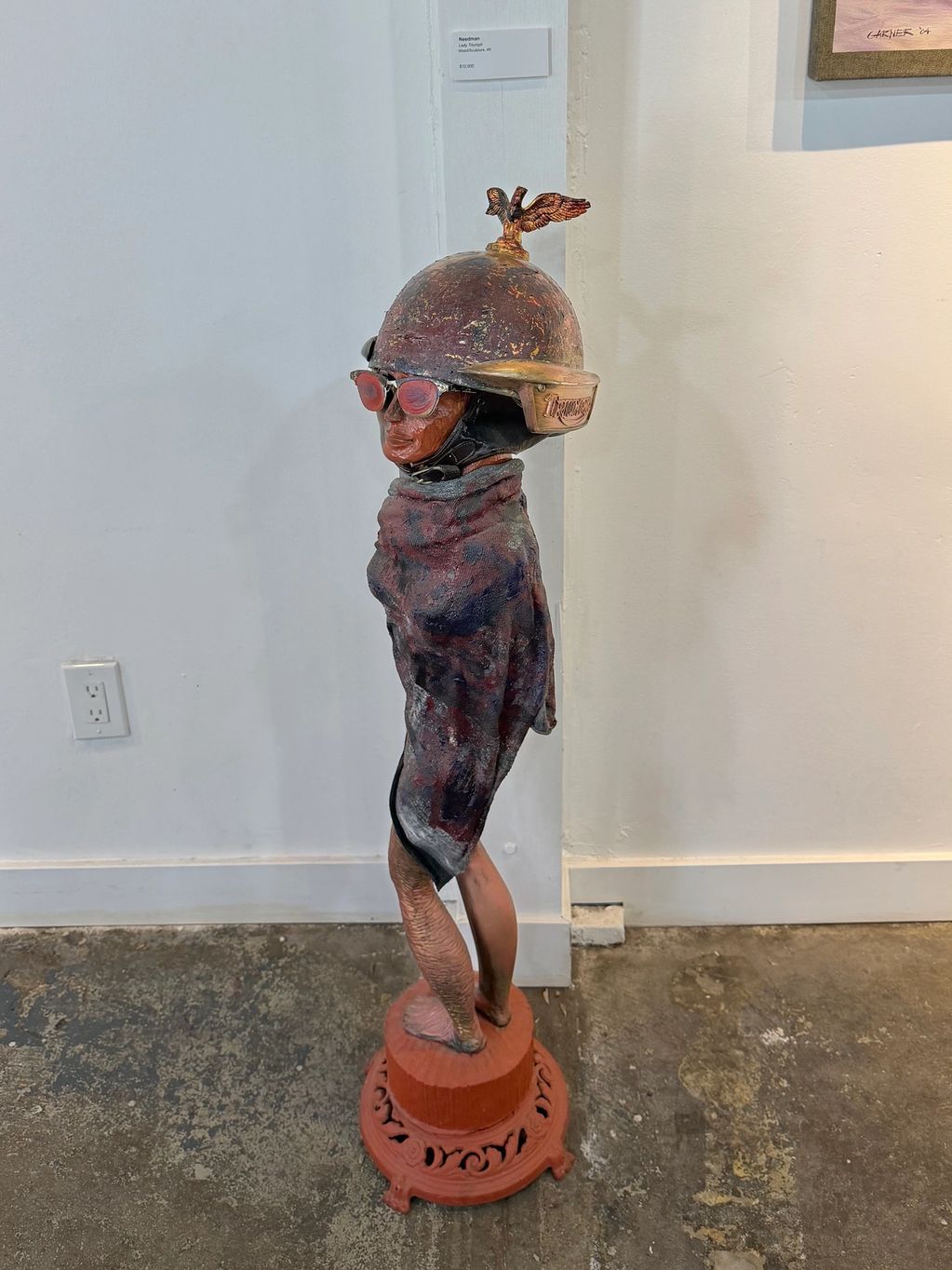 Sculpture of a figure wearing a helmet with wings and red glasses, draped in a textured cloth.