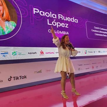 Conferencista Paola Rueda Lopez en tarima con backing grande atrás que dice su nombre y Colombia