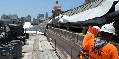 Two construction workers on a rooftop with city skyline in background.