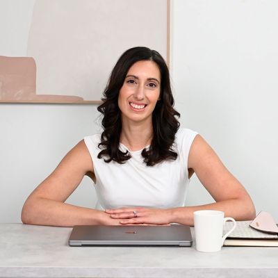 Smiling woman seated at a desk with laptop, coffee mug, and notebook.