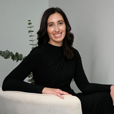 Woman in black dress sitting on a white chair, smiling.