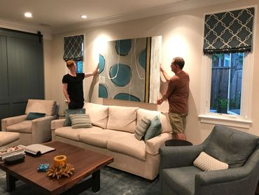 Two men hanging a blue and beige abstract painting in a cozy living room.
