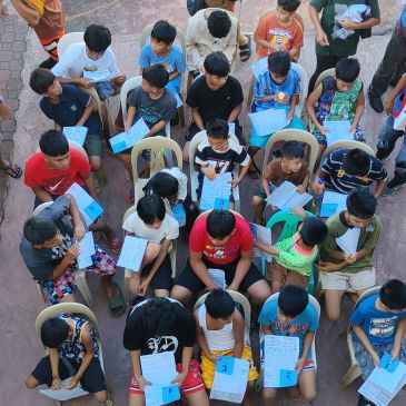 Children seated outdoors, holding papers and writing, engaged in an activity.
