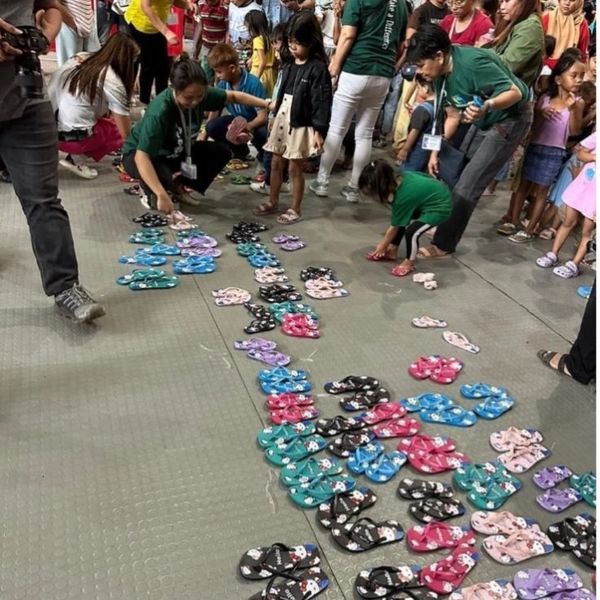 People distributing colorful slippers to children in a crowded indoor space.