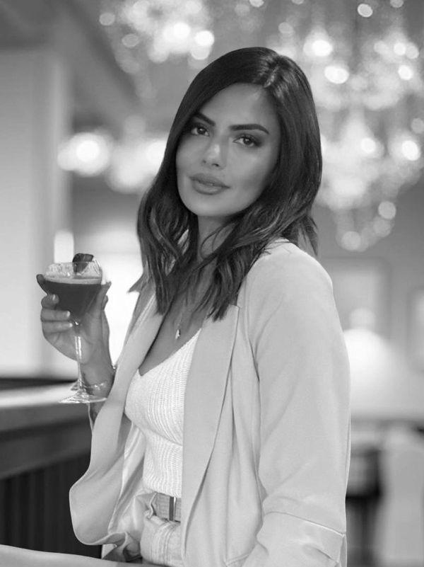 Woman in a blazer holding a red cocktail, sitting on a bar stool.