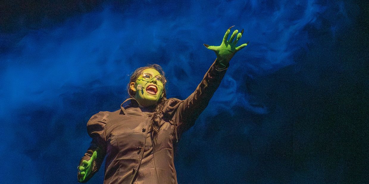 Actor in green makeup performs dramatically against a blue smoky background.